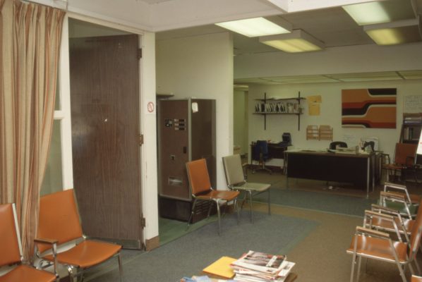 Intérieur de bureau avec décor d’époque, incluant chaises de salle d’attente, distributrice et bureau de travail. Des lumières fluorescentes de plafond illuminent la pièce.