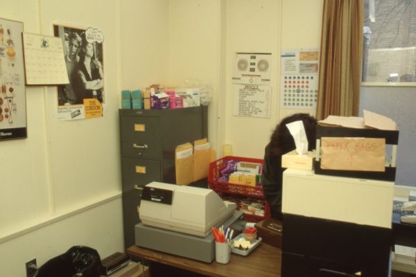 Bureau encombré avec classeur, caisse, affiches de contraceptifs et bureau de travail couvert de papiers et de fournitures.