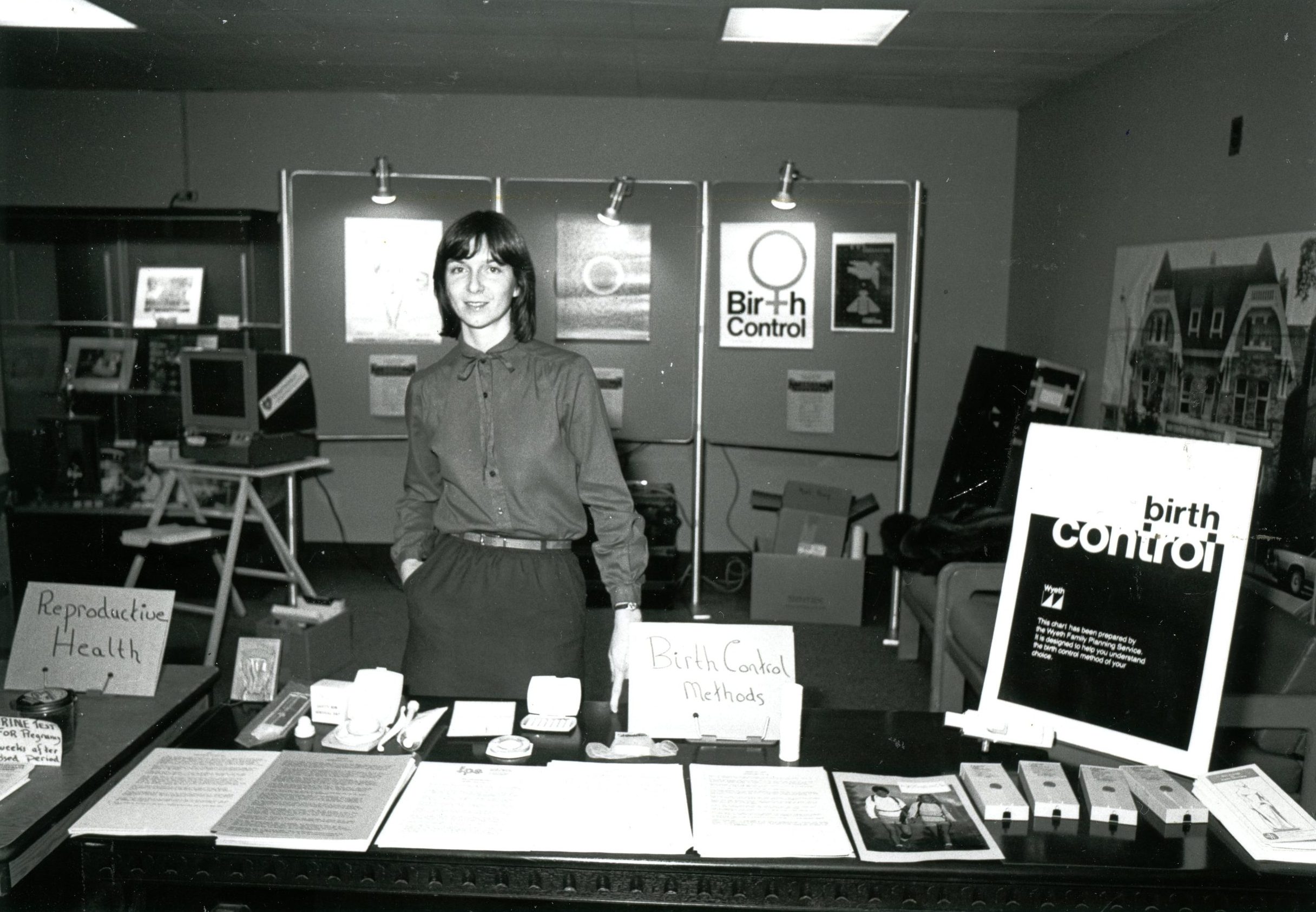 Une femme est debout derrière une table, souriant vers l’appareil photo. La table est couverte de matériel d’information et d’articles divers. Des enseignes sur la table et le mur se lisent : Reproductive Health and Birth Control (Santé reproductive et contraception). 