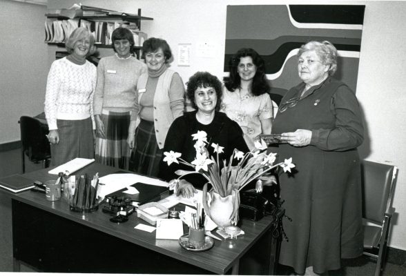 Six femmes dans un bureau. Elles sont réunies autour d’un bureau de travail en bois. Une femme est assise derrière le bureau, alors que les autres restent debout près d’elle. Le bureau est encombré de matériel de bureau, de stylos, de papiers et d’un vase de fleurs.