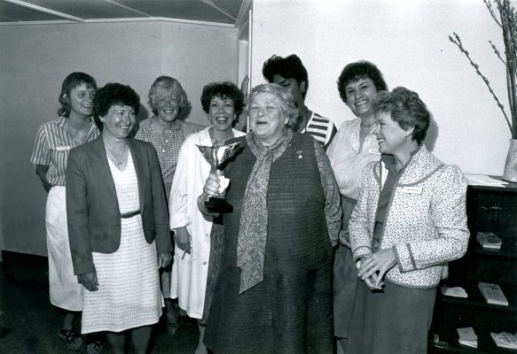 A group of eight women gathered in a room. The central figure, an elderly woman, is holding a large trophy and wearing a patterned scarf and dress.
