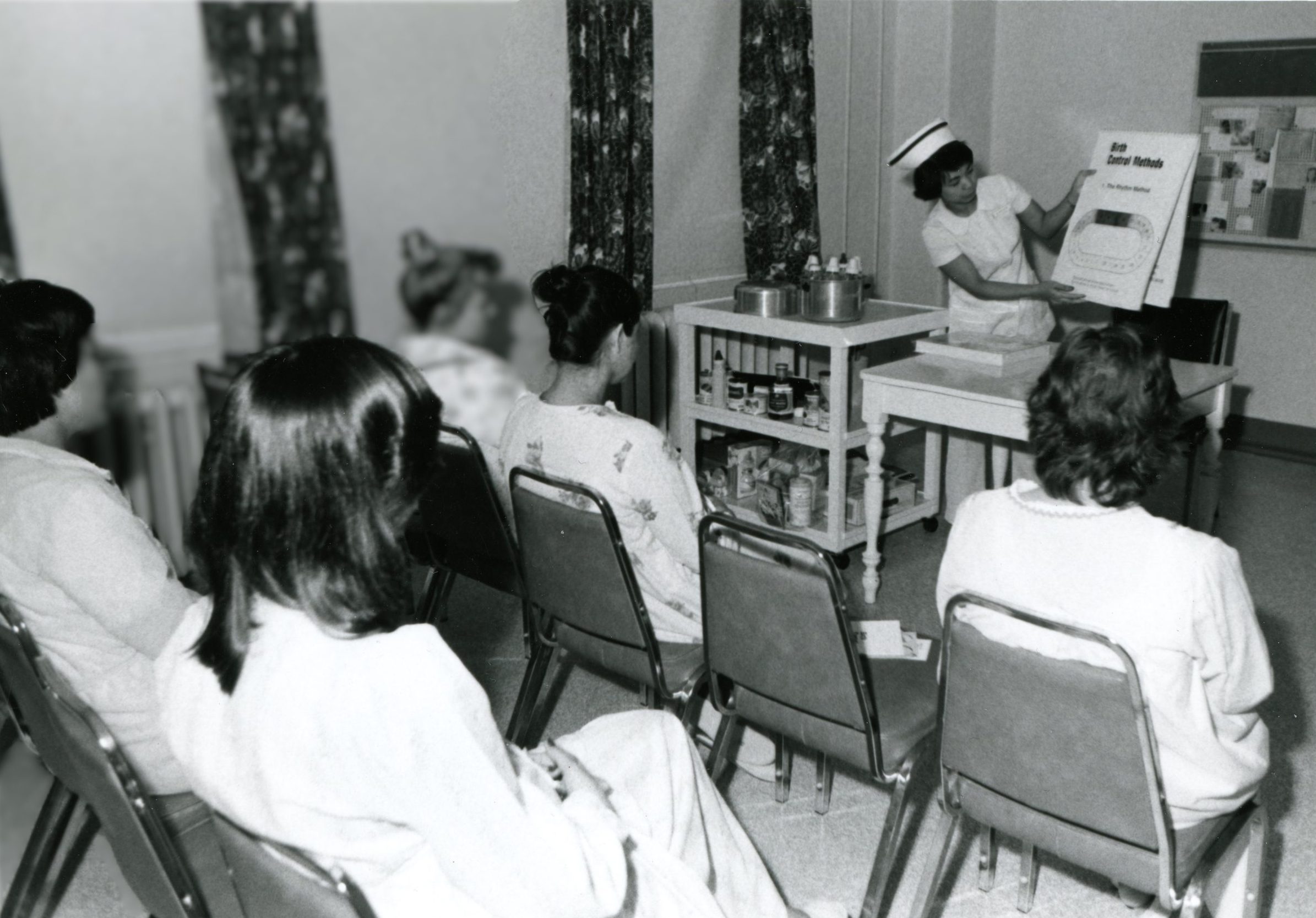 Un petit groupe de femmes en robes d’intérieur sont assises en rangées configurées comme une salle de classe. Elles font face à une infirmière debout devant elles. L’infirmière porte un uniforme traditionnel avec coiffe et leur enseigne à côté d’un chevalet. 