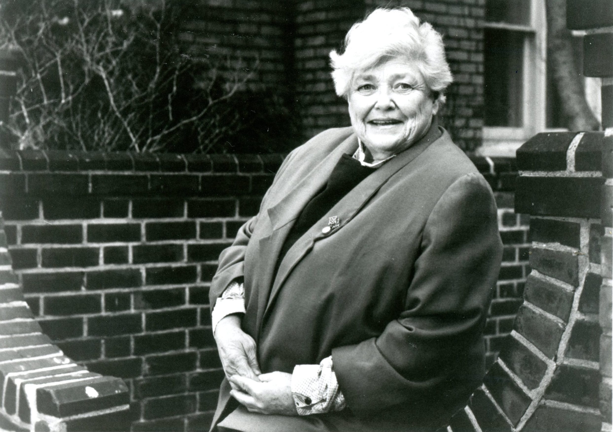 Une femme d’âge mûr aux cheveux clairs sourit à l’appareil photo. Elle porte un veston de couleur foncée sur un chemisier à col. Elle est positionnée devant un mur de briques qui forme une structure courbée ressemblant à un banc. 
