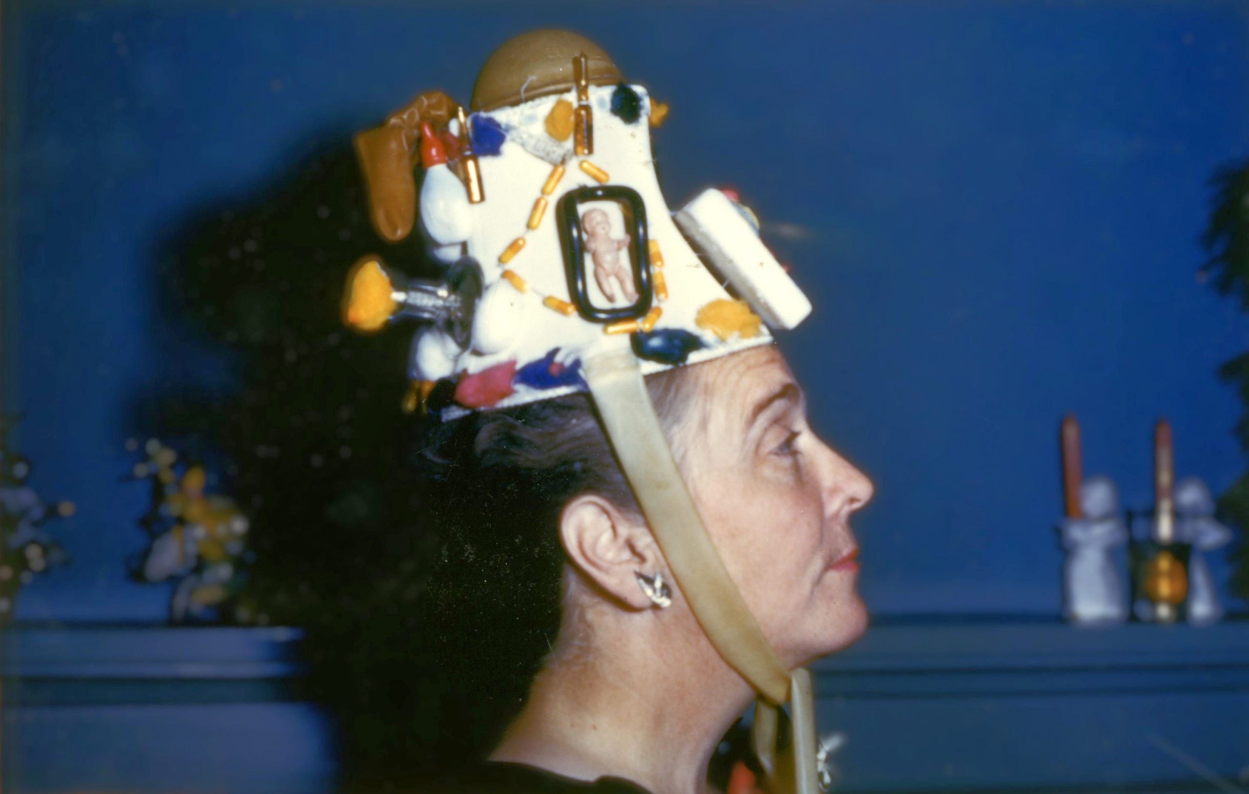 Une femme d’âge mûr de profil portant une parure de tête colorée faite de divers contraceptifs sur un fond bleu.