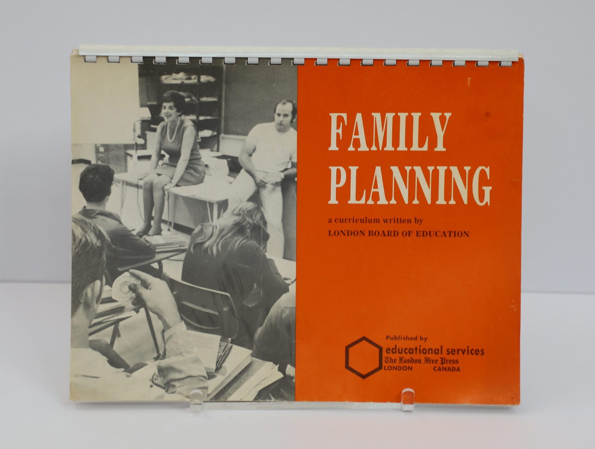 Petit livre éducatif rectangulaire avec reliure spirale le long du bord supérieur. La moitié gauche de la couverture présente une photo en noir et blanc révélant une scène de salle de classe. La moitié droite est un bloc orange sur lequel apparaît un texte en blanc se lisant : PLANIFICATION FAMILIALE, en anglais.