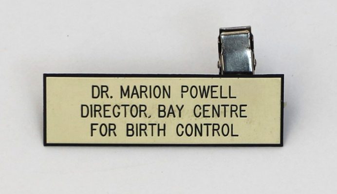 Porte-nom rectangulaire de couleur claire avec épingle au dos et texte en noir se lisant : DR. MARION POWELL, DIRECTOR, BAY CENTRE FOR BIRTH CONTROL.