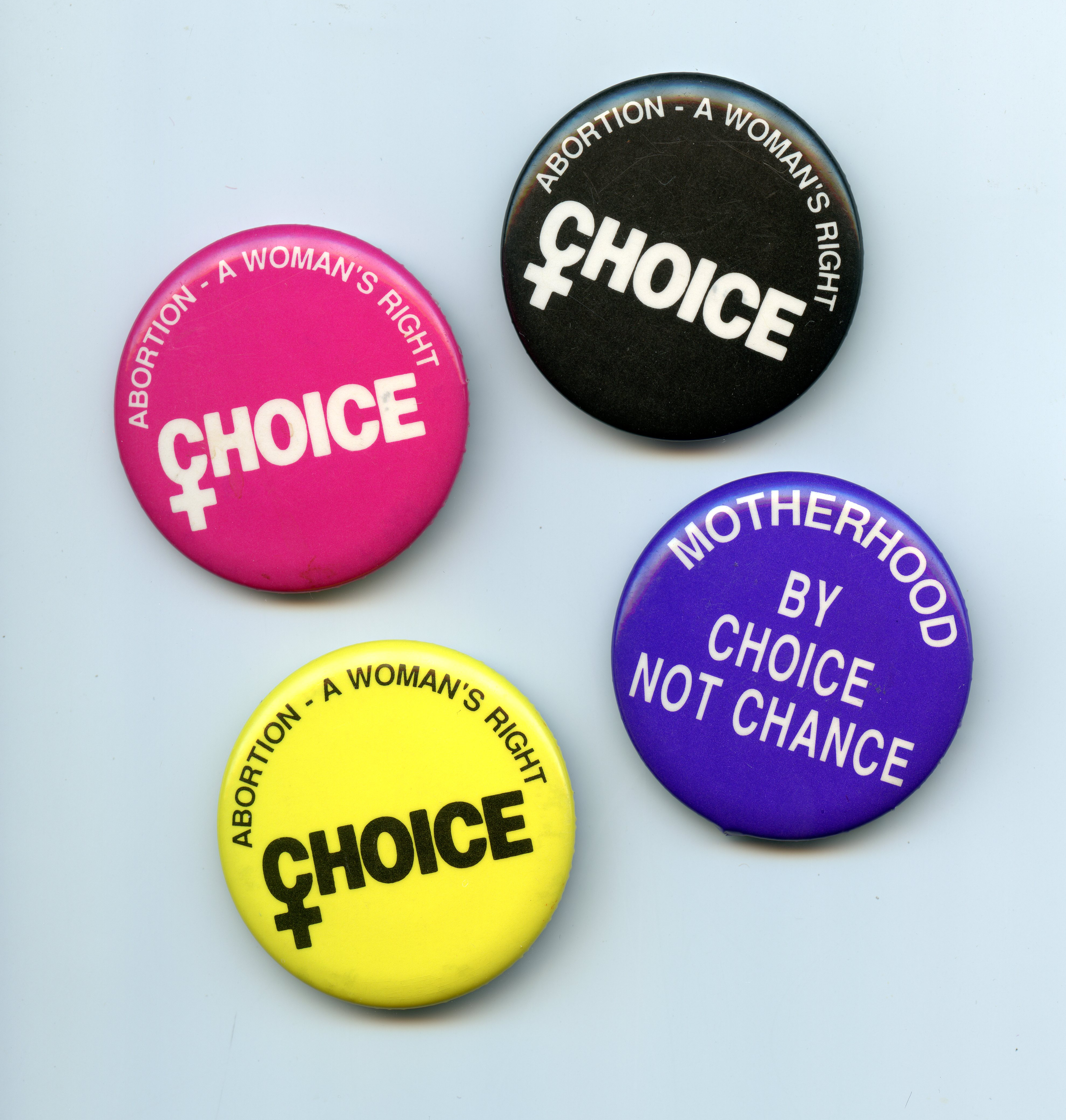 Four round buttons with slogans supporting women's reproductive rights on a white background. The top button is purple with white text stating Motherhood by Choice Not Chance and includes a small symbol that combines a female gender symbol with an equal sign. The bottom three buttons are pink, yellow, and black. These buttons have text that reads Abortion - A Woman's Right - CHOICE.
