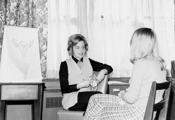 Deux femmes assises dans une pièce se parlent; l’une d’entre elles tient un modèle et l’on voit une illustration anatomique de l’appareil reproducteur féminin sur un chevalet. Un radiateur se trouve sous une fenêtre ornée de rideaux floraux.