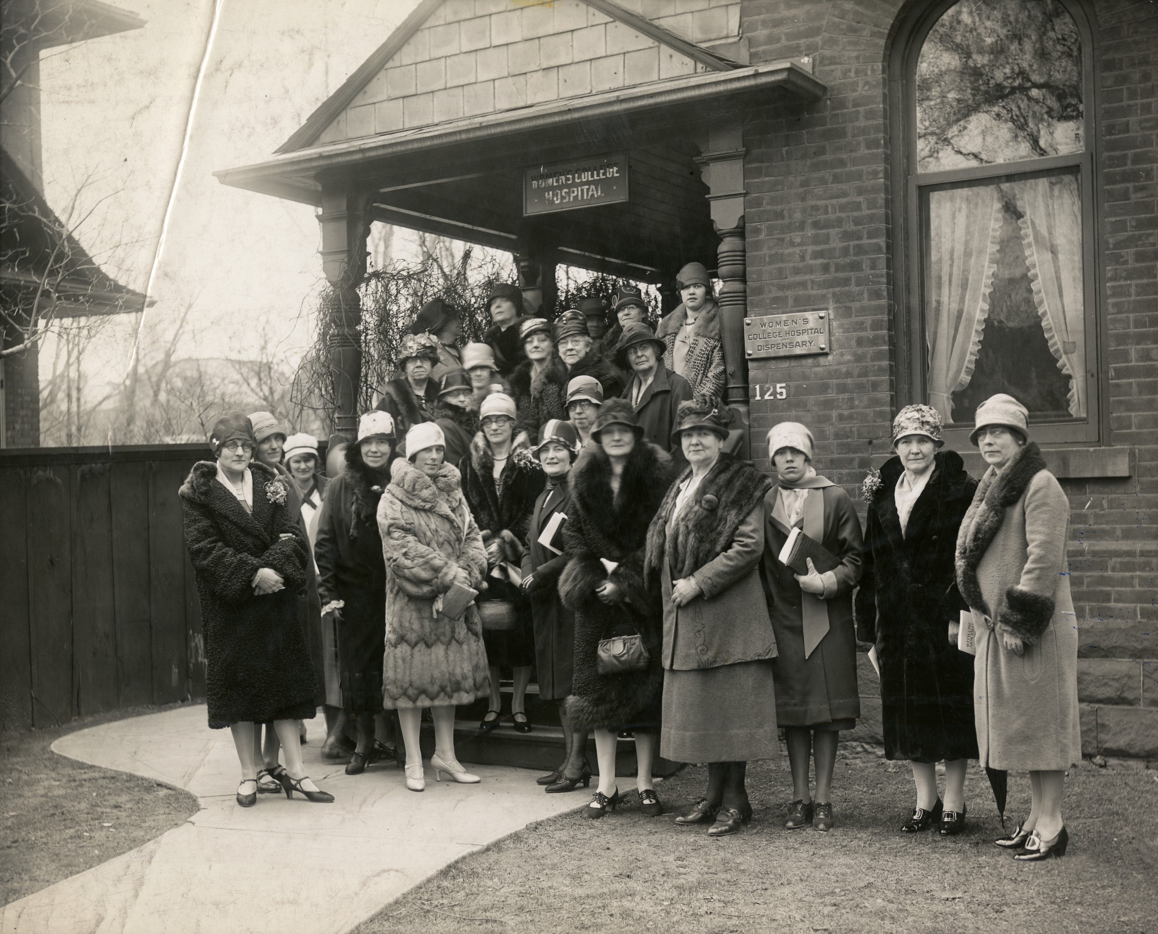 Photo en noir et blanc montrant un groupe d’environ 25 femmes posant devant un immeuble en briques avec une véranda. Les femmes sont vêtues à la mode des années 1920, portant des manteaux et des chapeaux cloches.