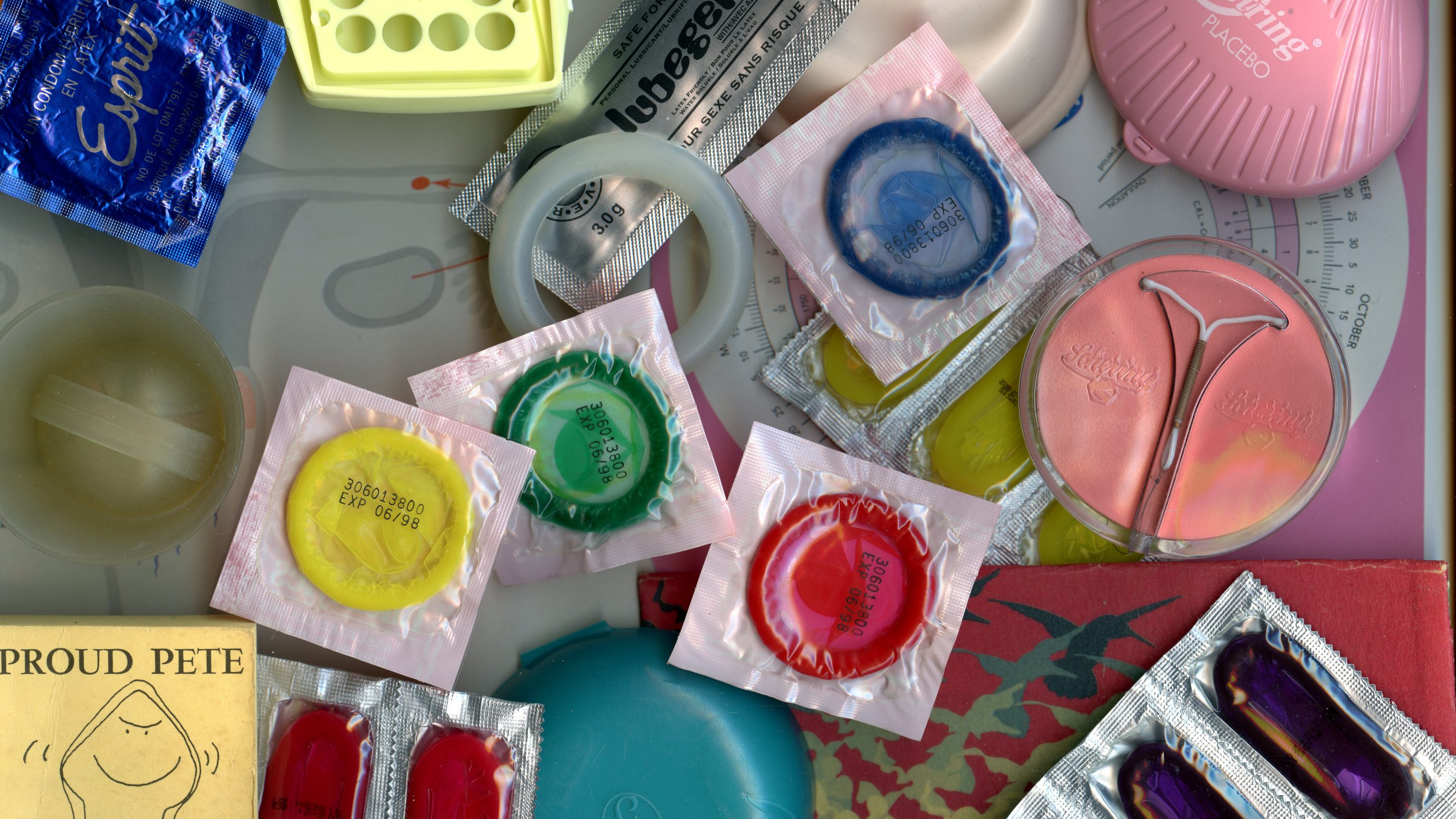 A collection of vintage birth control products arranged neatly on a flat surface. The items include pill packets, contraceptive sponges, condoms, and product packaging with diverse designs. Colors range from bright red, pink, and yellow to understated off-white. Each product features labeling or branding text. Collection de produits contraceptifs d’époque disposés avec soin sur une surface plane. Parmi les articles figurent des boîtes de pilules, des éponges contraceptives, des condoms, et des emballages de produits aux motifs divers. Les couleurs vont du rouge vif au blanc cassé discret en passant par le rose et le jaune. Chaque produit porte une étiquette ou un nom de marque.