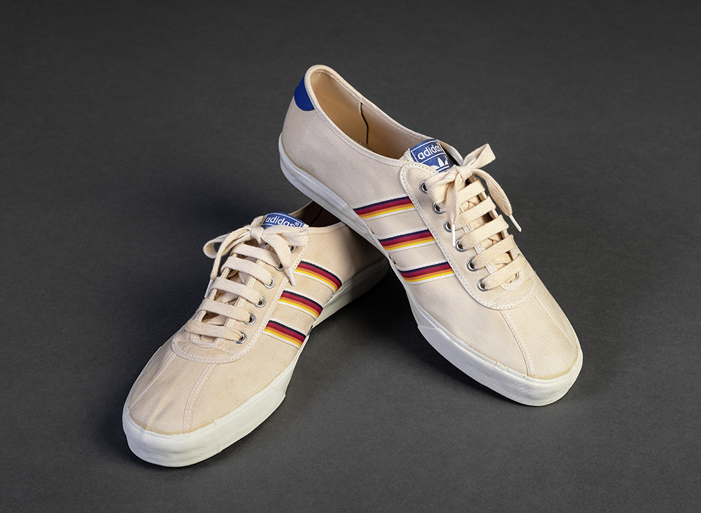 Deux chaussures de toile beige, aux lacets de même couleur, forment une paire. Trois bandes rayées multicolores décorent les côtés, tandis que sur les languettes, le logo trifolié d’adidas apparaît en blanc sur fond bleu.