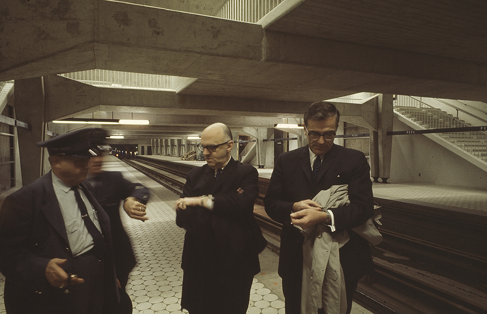 Sur le quai de la station de métro Peel, le maire Jean Drapeau et le président du comité exécutif de Montréal, Lucien Saulnier, consultent leurs montres. Deux agents en uniforme les accompagnent. L’un d’eux regarde aussi sa montre.