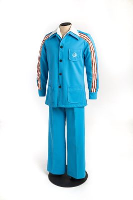 A mannequin is dressed in the photographer uniform: straight-leg blue trousers and a jacket in the same colour, buttoned over a white shirt. A striped ribbon runs down the jacket sleeves and the breast pocket is embroidered with the Games logo.