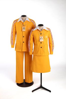 Deux mannequins de couture présentent des uniformes oranges. À gauche, l'uniforme pour hommes : un pantalon droit à plis et une veste, aux manches ornées d’un liseré tricolore. À droite, l'uniforme pour femmes : une jupe mi-longue et une veste, ouverte sur une chemise blanche. Boutonnées sur une chemise blanche, les vestes portent le logo blanc des Jeux brodé sur leurs poches. Au cou des mannequins se trouvent des cartes d’employés plastifiées.