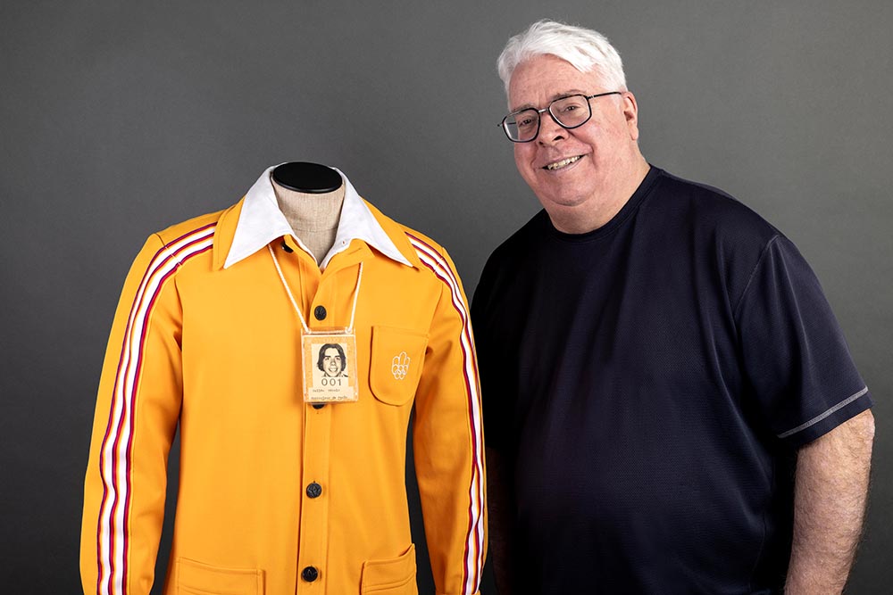 Gérald Vallée se tient près d’un mannequin de couture présentant son uniforme orange des Jeux olympiques de 1976. Boutonnée sur une chemise blanche, la veste orange porte le logo blanc des Jeux brodé sur sa poche. Au cou du mannequin se trouve une carte d’employé plastifiée.
