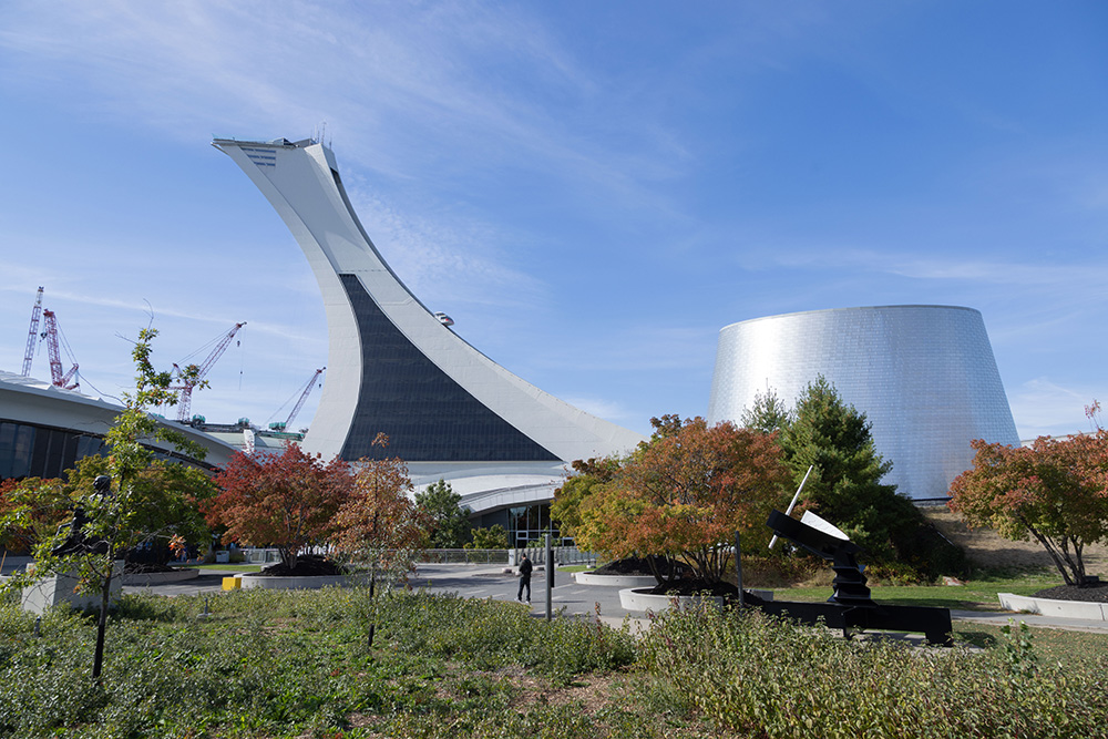 À côté de la tour inclinée du Stade olympique de Montréal, le Planétarium se distingue par sa façade en aluminium et sa structure en forme de cône tronqué. Une personne se promène sur une allée bordée d’espaces verts et d’arbres.