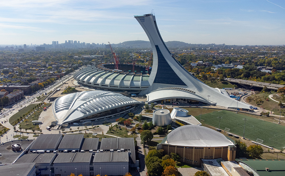 Vus du ciel, le Stade olympique et le Biodôme s’imposent devant Montréal. Trois grues dépassent du toit ouvert du stade. Au loin se distinguent le centre-ville et le mont Royal.
