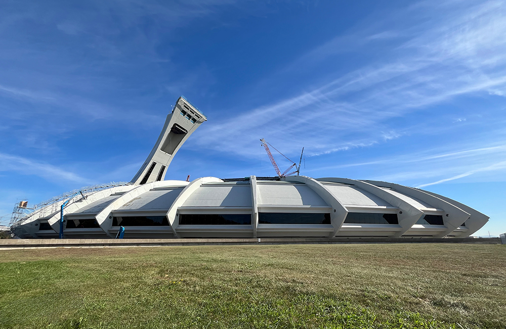 Le Stade olympique et sa tour inclinée se dressent entre un ciel bleu et un terrain gazonné. Deux grues dépassent du dôme du stade, alors qu’à gauche, un échafaudage métallique complexe en garantit l’accès.