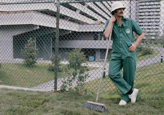 Devant la clôture des habitations du Village olympique, un employé de l’entretien prend la pose, appuyé sur un balai-brosse. Coiffé d’un chapeau cloche blanc, l’homme moustachu porte une combinaison vert émeraude à la poche brodée du logo des Jeux.