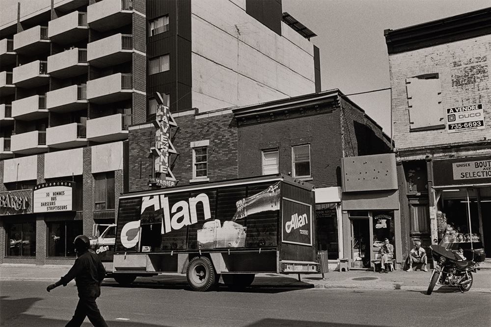 En noir et blanc, un camion de livraison de la marque de boissons gazeuses Allan circule sur la rue Sainte-Catherine, devant une taverne et un cabaret de danseurs nus. Un passant marche et deux hommes sont assis, près d’une cordonnerie et d’une moto stationnée.
