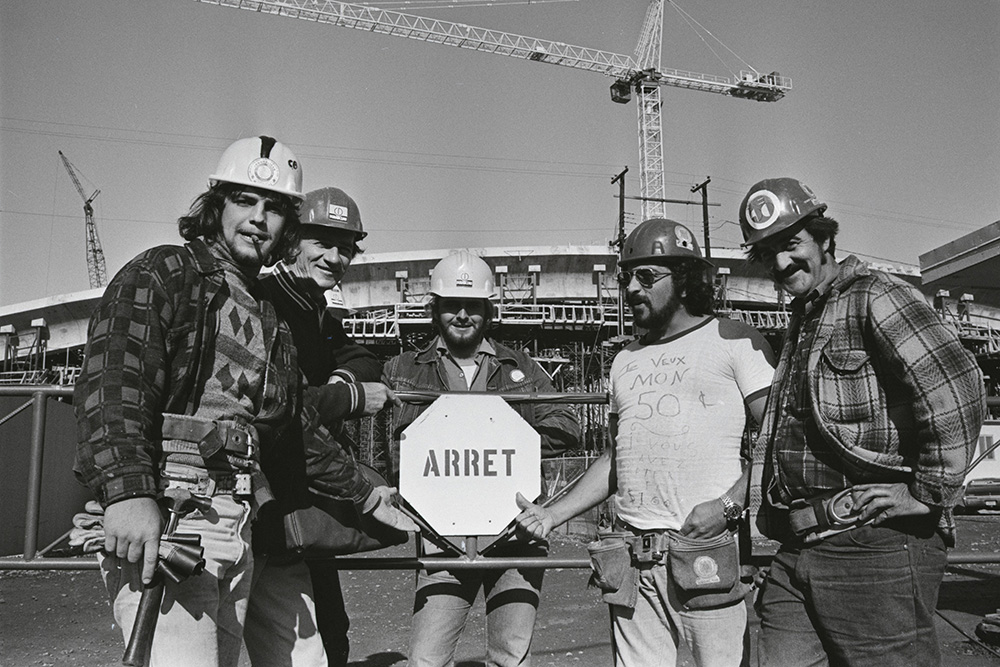 En noir et blanc, cinq travailleurs coiffés de casques de chantier entourent un panneau d’arrêt devant le Vélodrome en construction. Sur le t-shirt de l’un d’eux est notamment écrit : Je veux mon 50 cents.