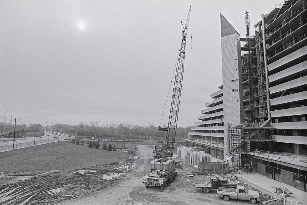En noir et blanc, une grue se dresse près du Village olympique inachevé. Des matériaux de construction s’entassent au bas d’un bâtiment pyramidal et une chute à débris relie le sommet à un conteneur.
