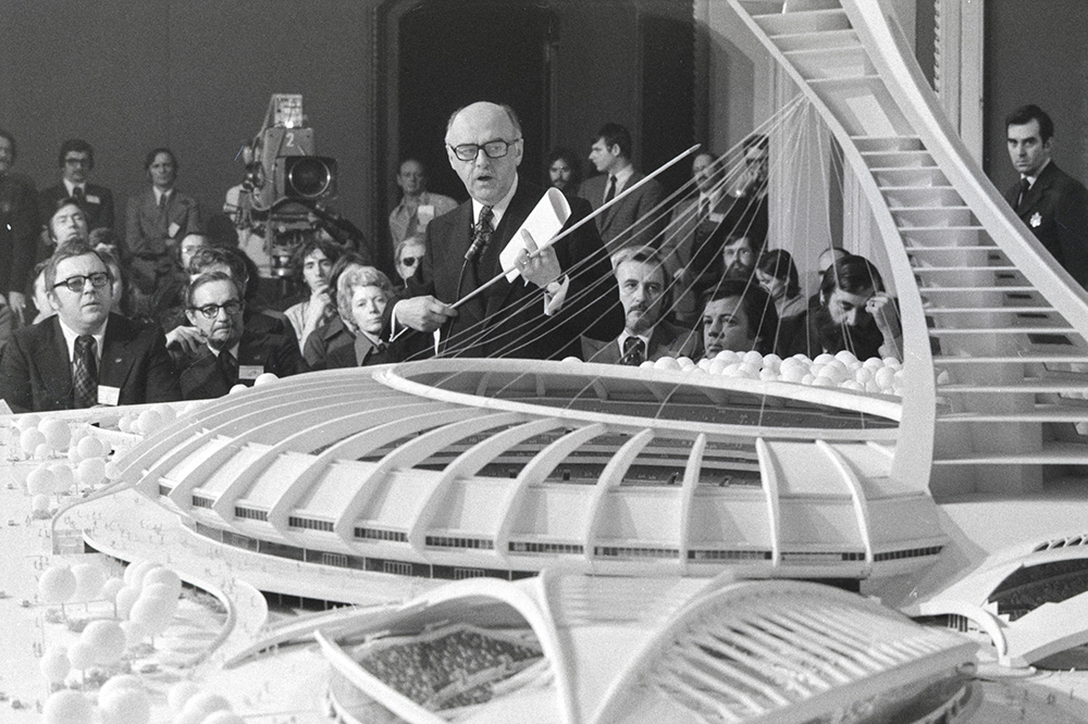 En noir et blanc, Jean Drapeau présente une maquette du Stade olympique et du Vélodrome à une vingtaine de membres de la presse. Le maire tient un document plié et un pointeur télescopique entre ses mains.
