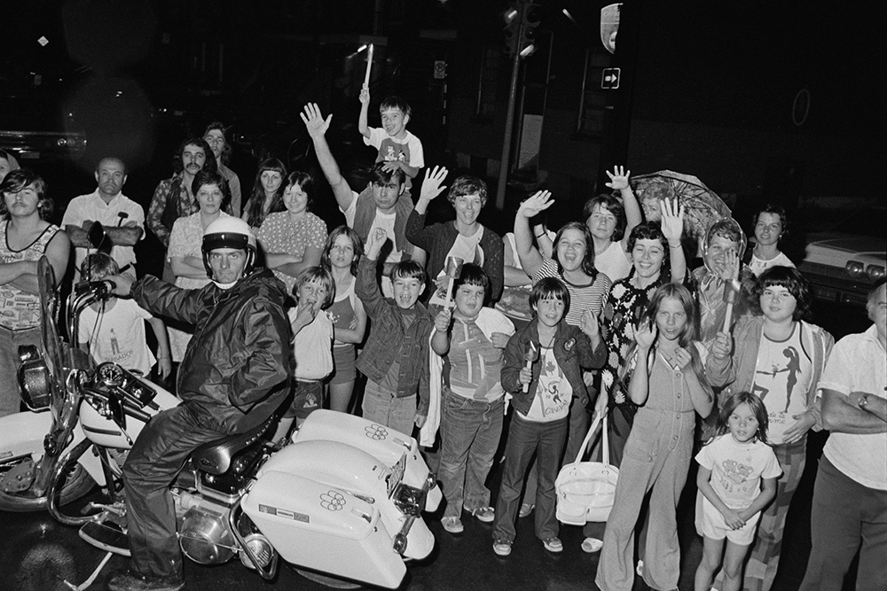 En noir et blanc, une foule composée de familles attend le passage de la flamme olympique. Plusieurs personnes saluent, souriantes. D’autres attendent, bras croisés. Un homme est assis sur une moto blanche portant le logo des Jeux de Montréal.