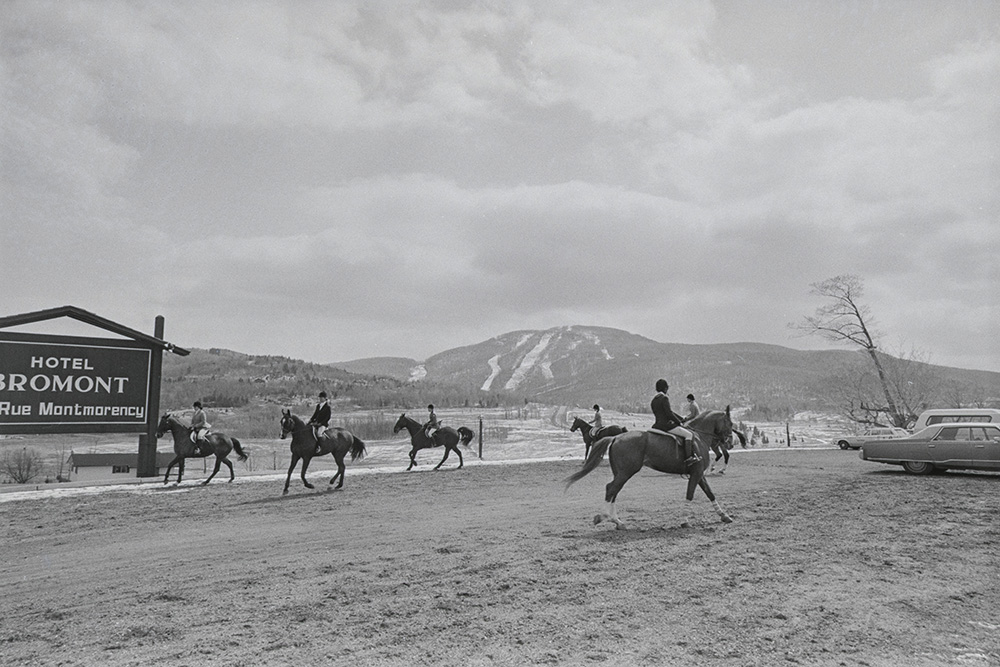 En noir et blanc, six athlètes en tenue d’équitation montent à cheval sur un terrain dégagé. Une montagne de ski se dresse au loin. Près du groupe, un panneau annonce : Hôtel Bromont, rue Montmorency.