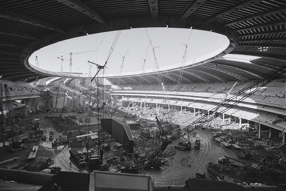 En noir et blanc, de nombreux engins de chantier et des matériaux s’entassent sur le sol argileux à l’intérieur du Stade olympique en construction. Des grues s’élèvent à travers son dôme, percé d’un orifice circulaire.