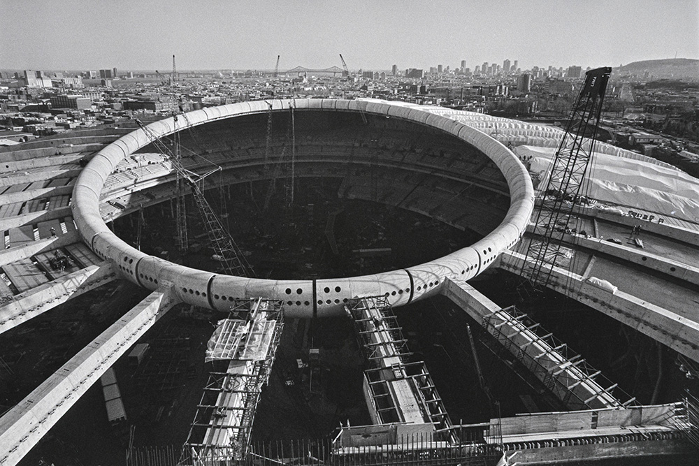 En noir et blanc, vue du ciel, une large ouverture cerclée d’un anneau perce le dôme du Stade olympique en construction. Des engins et des véhicules de chantier occupent l’intérieur du bâtiment.