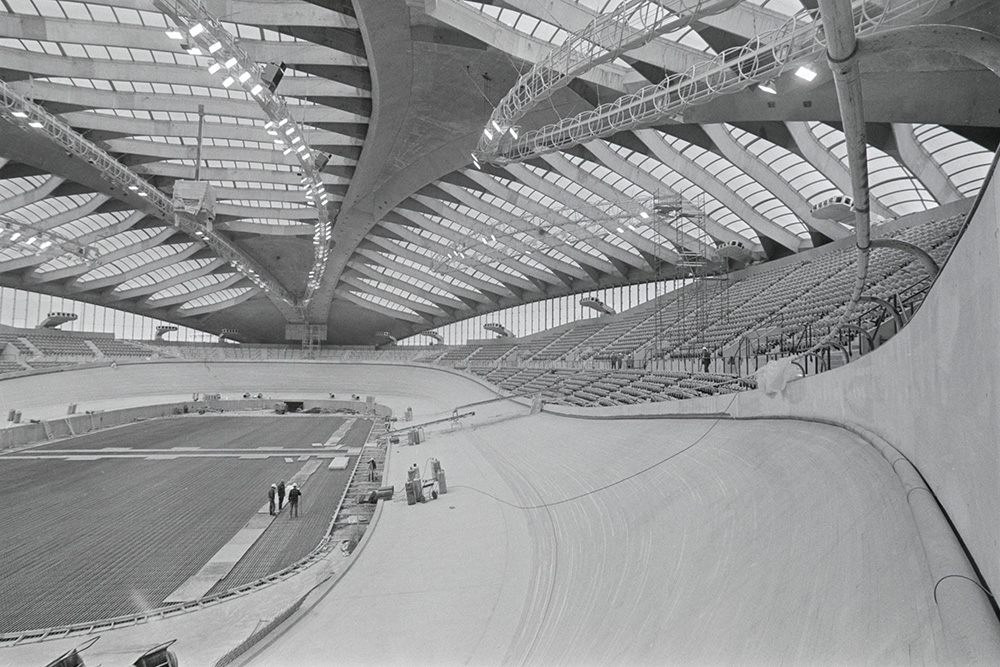En noir et blanc, l’intérieur du Vélodrome en construction présente une piste ovale aux tournants inclinés, des gradins et un toit à la structure de béton ajourée. Quatre ouvriers se trouvent dans l’espace dégagé au centre de la piste.
