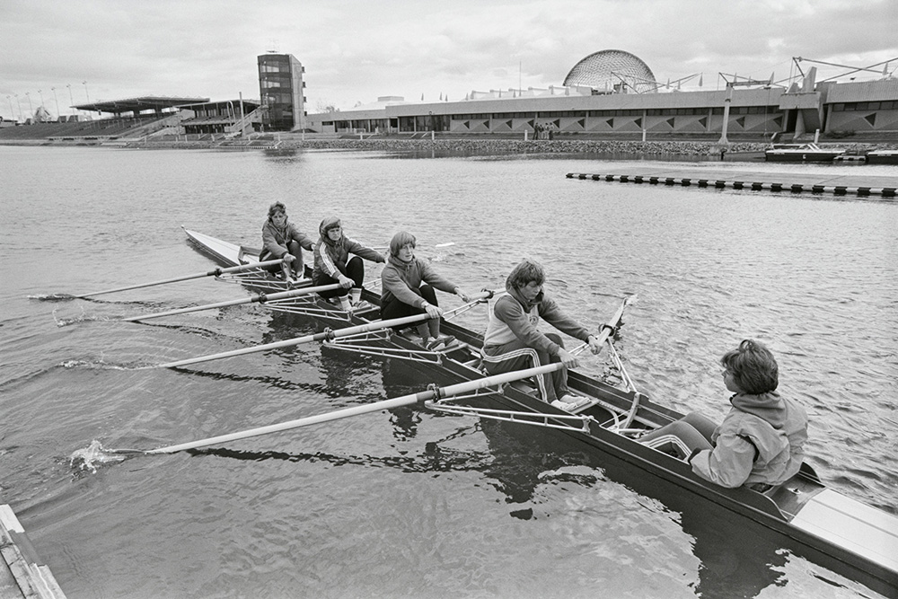 En noir et blanc, quatre femmes rament à bord de leur aviron, alors qu’une cinquième, la barreuse, leur fait face. Loin derrière, des gradins et la grande structure sphérique du pavillon des États-Unis d’Expo 67 se dressent sous un ciel voilé.