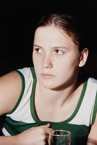 Gros plan sur le visage et les épaules de la nageuse canadienne Anne Jardin, alors âgée de 17 ans. Ses cheveux bruns attachés, elle porte un débardeur vert et blanc et affiche un air sérieux. Un verre transparent repose devant elle.