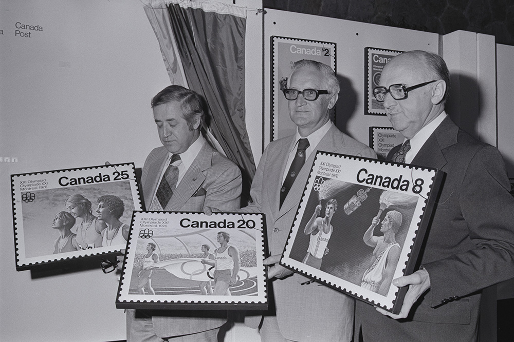 En noir et blanc, le maire Jean Drapeau, le commissaire général des Jeux olympiques, Roger Rousseau, et le ministre fédéral des Postes Bryce Mackasey présentent trois timbres olympiques, en grand format.