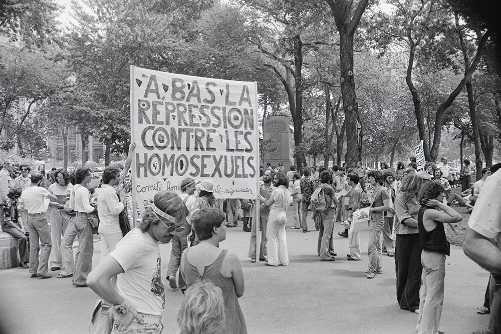 En noir et blanc, plus d’une cinquantaine de protestataires, jeunes pour la plupart, sont réunis au square Dorchester. Sur une grande affiche tenue par deux personnes, il est écrit : À bas la répression contre les homosexuels.