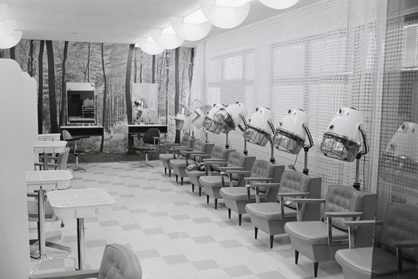 En noir et blanc, huit fauteuils-séchoirs s’alignent dans un salon de coiffure désert au sol en damier. Deux miroirs sont fixés au mur du fond, couvert d’un papier peint représentant une forêt.