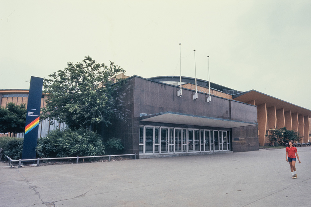 La vaste entrée vitrée du centre Pierre-Charbonneau est surmontée d’une façade en pierre calcaire grise. Le reste du grand bâtiment est couvert de briques orangées. Devant le centre sportif, une jeune femme habillée en short bleu et t-shirt rouge se promène.