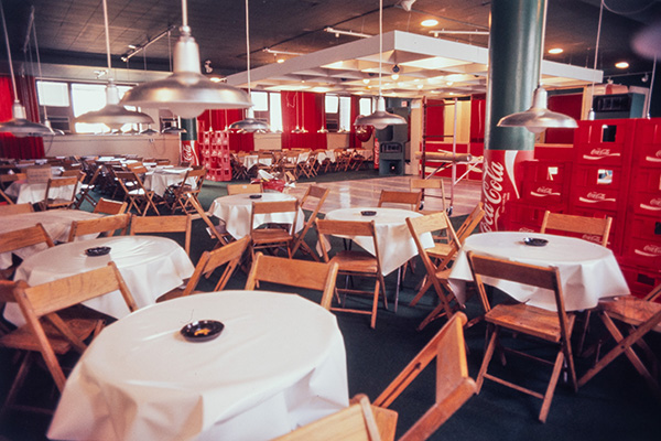 Dans la discothèque, de nombreuses petites tables rondes et des chaises en bois sont installées autour d’un plancher de danse. Des cendriers occupent le centre des nappes blanches et de nombreuses caisses en plastique rouge de la marque Coca-Cola sont entreposées.