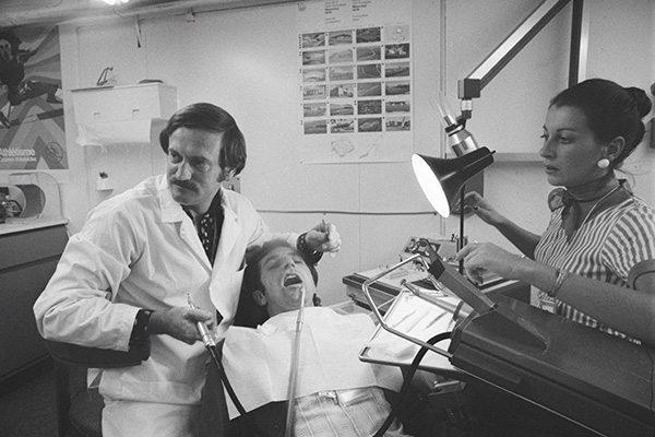 En noir et blanc, un dentiste tient un instrument dentaire auprès d’un homme allongé, la bouche ouverte, sous l’éclairage vif d’une lampe. Le dentiste et une femme qui l'assiste regardent ailleurs, dans la même direction, l’air concentré.