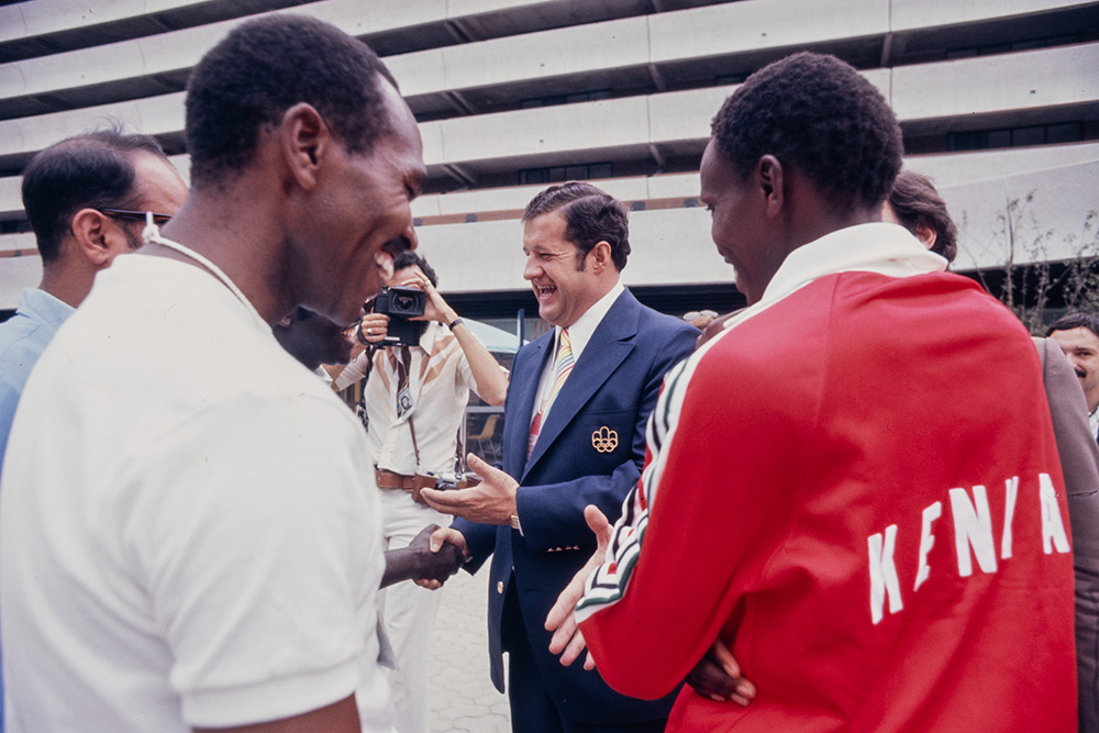 Le directeur général et maire du Village olympique, Yvan Dubois, discute avec des athlètes kényans vêtus d’une tenue principalement rouge et blanche. Debout devant le Village olympique, le maire, habillé d’un complet sombre, et les sportifs se sourient.
