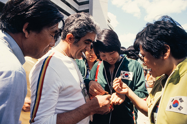 Un petit groupe d’athlètes portant des vêtements verts décorés d'un écusson représentant le drapeau de la Corée du Sud échangent des épinglettes, avec un homme qui fume.