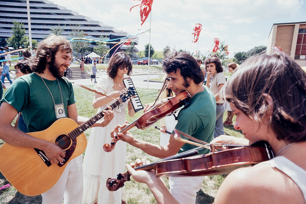 Quatre musiciennes et musiciens jouent devant le Village olympique de Montréal. Ils se tiennent debout en cercle sur le gazon. L’un joue de la guitare, un homme et une femme jouent du violon, et une quatrième musicienne joue du piano à bouche.