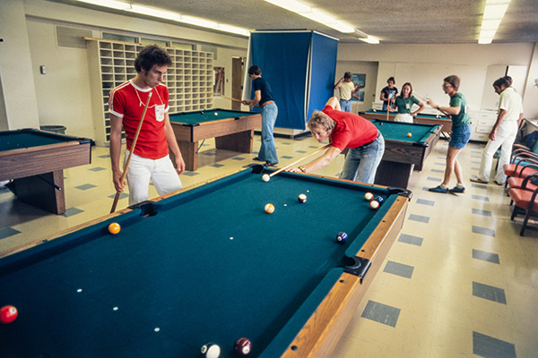 Dans une salle du Village olympique de Montréal, quelques personnes habillées de manière décontractée jouent au billard. Cinq tables sont réparties dans la pièce aux murs jaune pâle. Plusieurs parties sont en cours.
