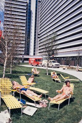 Devant les grands bâtiments du Village olympique, cinq hommes et femmes en maillots de bain sont allongés sur des chaises longues jaunes installées sur du gazon. Certains sont assoupis, d’autres ont un livre à la main.