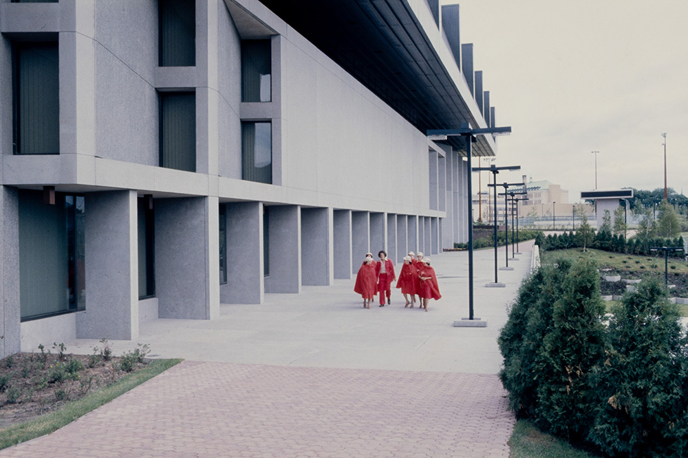 Devant le grand complexe sportif Claude-Robillard en béton gris clair, un groupe de guides et d'hôtesses des Jeux se déplacent. Ils portent des uniformes unis rouges.