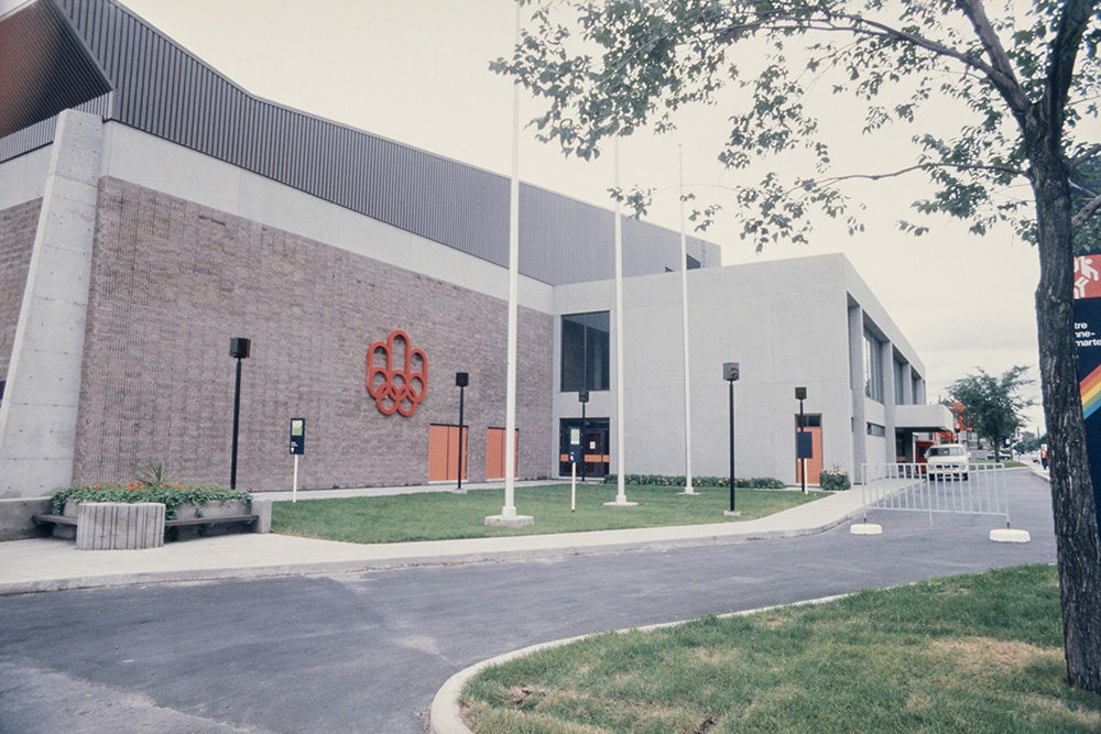 Le grand bâtiment en béton du centre Étienne-Desmarteau borde une allée asphaltée et des terre-pleins couverts de gazon. Le logo rouge des Jeux olympiques de Montréal apparaît en grand sur un mur de l’édifice.