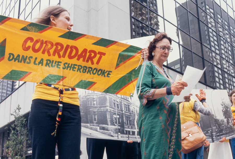 Dans Montréal, une femme brandit une banderole où est écrit : Corridart dans la rue Sherbrooke. Une autre fixe du regard une feuille de papier entre ses mains. D’autres portent des photographies grand format en noir et blanc de bâtiments historiques.