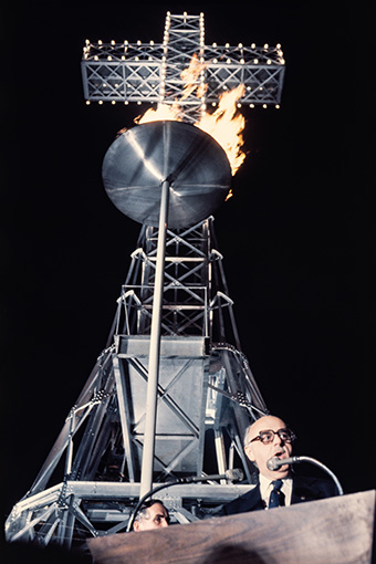 De nuit, le maire de Montréal Jean Drapeau prononce un discours. Derrière lui, la croix du mont Royal domine la flamme olympique qui brûle dans une haute vasque en métal.