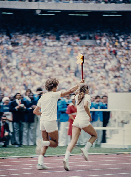 Stéphane Préfontaine et Sandra Henderson courent, de dos, sur la piste du Stade olympique de Montréal. Vêtus de shorts et t-shirts blancs, les deux jeunes athlètes brandissent ensemble la torche rouge enflammée, dont la partie supérieure est noire.